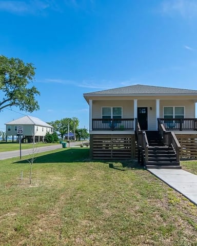 Elevated Beach House With Front Porch And Dark Wooden Stairs, Surrounded By Grass And Mature Oak Tree, Neighboring Coastal Homes Visible.