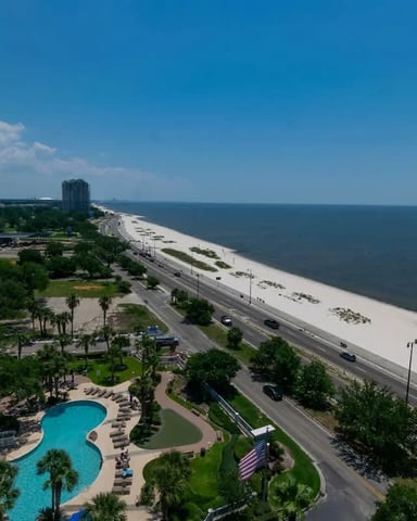Elevated View Of Beachfront Resort With Pool, Palm Trees, Sandy Beach, Coastal Highway, And Gulf Waters Under Clear Blue Sky